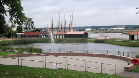 Geograph/John Firth The Maldon amphitheatre in front of a body of water, pedestrians passing by, in the background is an estuary and tall flags