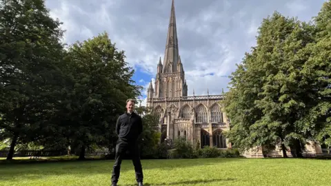 St Mary Redcliffe Brutus is standing on a lawn in front of St Mary Redcliffe, with the church in the background and trees either side. He is wearing all black with his hands behind his back and his feet apart. The sky is blue with lots of grey cloud.