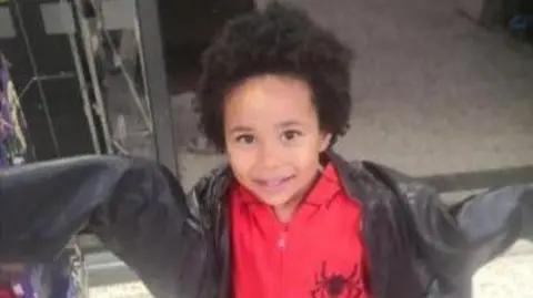 Wearing an oversized black coat and a red jacket with a spider motif, four-year-old Kobi Macharia Dooly looks directly at the camera. 