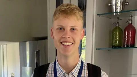 Family photo Sebastian is smiling looking at the camera - he is in a kitchen, wearing a check shirt and a rucksack with a blue lanyard.