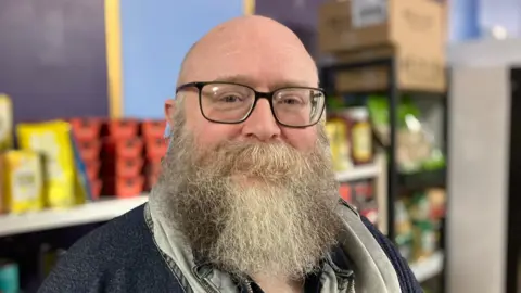 A bald man with a long grey beard and wearing glasses looks towards the camera. Behind him, out of focus, shelves filled with food items can be seen.