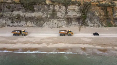 BCP Two yellow lorries on a beach and a smaller black vehicle. The picture was taken from a bird eye's view. It is an overcast day.