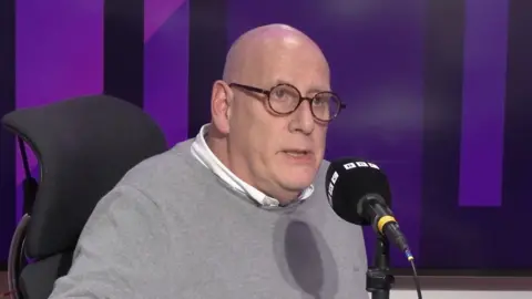 Bald headed man with glasses in a BBC radio studio. 