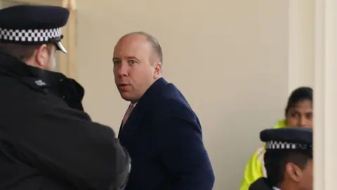 A picture of Matt Hancock, with a shaved head and wearing a blue overcoat, entering the Covid inquiry building in Paddington, west London. A policeman is looking on in the foreground.