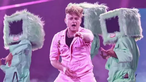 EPA Sam Battle aka Look Mum No Computer performing on stage during Eurovision dress rehearsals in a pink jumpsuit pointing towards the camera, with three dancers behind him wearing fur-lined computer monitors with red lips over their heads