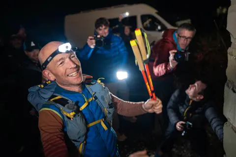 Kim Collison wearing a running light and carrying walking poles celebrates his Spine Race victory surrounded by photographers