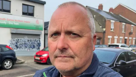 A close up image of a man with short grey hair. He is standing in front of a white building with a green banner and graffiti on the front of it. There is a row of cars parked behind him. 