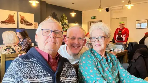 Juner Family Three people looking at the camera all wearing glasses. The man on the left is wearing a blue cardigan and red shirt, the man in the middle is wearing a white t-shirt and the woman on the right is wearing a green patterned blouse.