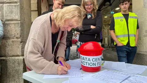 A woman writing her contact details on a piece of paper on a table with other papers and a small red bucket to collect money for the Data Centre Action Fund