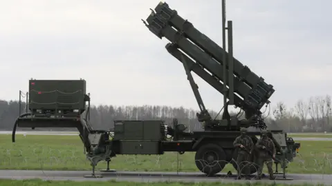 Two armed people in camouflage stand in front of a Patriot missile system