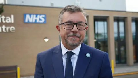 SHAUN WHITMORE/BBC Director of estates Paul Brooks stands outside the new diagnostics centre at the Queen Elizabeth Hospital in King's Lynn. He is wearing a blue suit and a navy tie. He is wearing black-framed glasses.