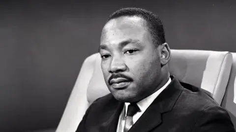 A black and white image shows Dr Martin Luther King sitting in a chair on a TV set. He is looking down, his eyes not meeting the camera. He has short hair and a neat moustache, and is wearing a dark jacket, tie and white shirt. 