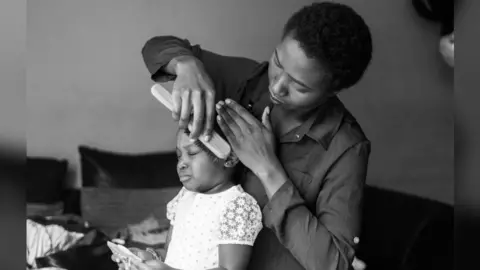 Iko Ojo Mercy /Eye Mama Project Iko Ojo Mercy is wearing a dark shirt and shown brushing her daughter's hair as her daughter shuts her eyes.