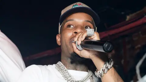 Getty Images Tory Lanez rapping at the Sorry For What Event on September 28, 2022 in New York City. He's wearing a chunky, expensive-looking silver bracelt and matching necklace. He's holding a microphone close to his face, and the side of a very large, diamond-studded ring on his pinky finger is visible.