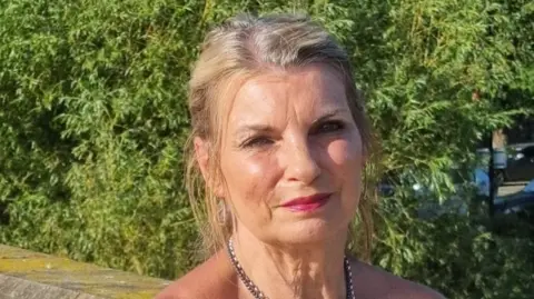 A woman with light coloured hair, wearing a necklace, is looking directly at the camera. The picture has been taken outside, against a wall and hedging