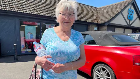 Margaret Smillie has shorty white hair and wears glasses. She is wearing a patterned white and blue top. She is holding a large bottle of evian water in her arms.