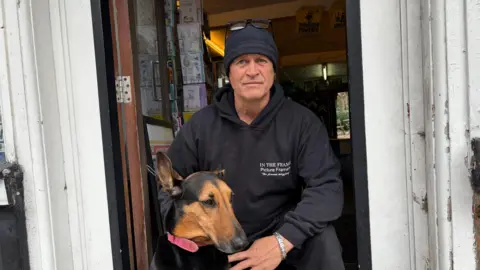 A man in a black hoodie, crouches in a doorway petting his dog.