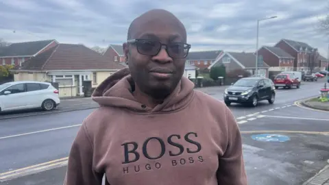 Adewale Akinade, who is bald, wearing a pink jumper with Hugo Boss branding and sunglasses. He is stood in front of Gors Avenue