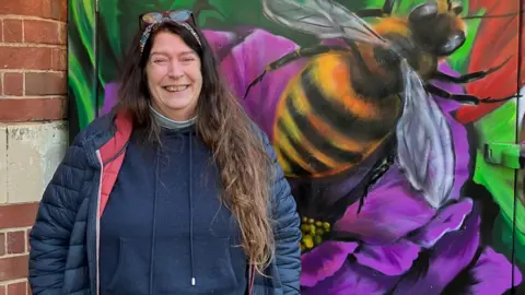 A woman with long brown hair and wearing a blue coat, blue jumper, headband and sunglasses stands in front of a colourful mural depicting a large bee.