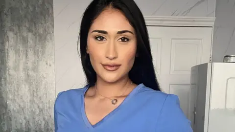 @healthwithnyrah Nyrah Saleem, looking at the camera and slightly smiling, she is wearing a blue pharmacist's uniform. The background has a white door.