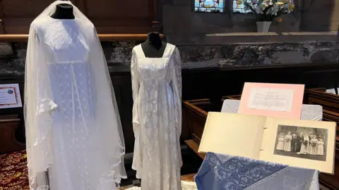 Andrew Rogers Two wedding dresses displayed on mannequins. The one to the left has a high neck and empire waistline, a pattern of stitched diagonal crosses and a long veil draped behind it. The other has a high waistline, a square neckline, long sleeves and stitched pattern of flowers. To the side an open photograph album resting on a table shows a wedding picture.