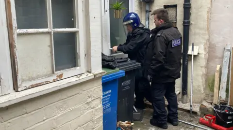 Two men wearing black clothing are standing in front of a white door in what looks like a small patio area. One is wearing a black coat with "POLICE" emblazoned on the back. The other, wearing a blue helmet with a face shield, is trying the door handle.