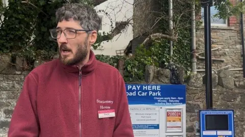 Handout Daniel Adams at Marson Road Car Park, wearing a burgundy zip up fleece with Stationery House written on it. He is standing beside a parking meter and pay here sign.