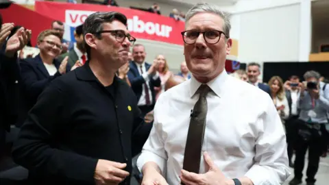 Starmer and Burnham pictured together at the launch of the Labour manifesto in 2024, surrounded by an applauding crowd.