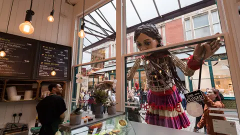 Getty/Anadolu Little Amal peaks through a bakery window to scope out a bite to eat