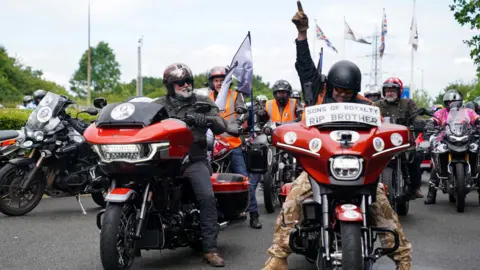 Hairy Biker Si King on his motorbike at the National Motorcycle Museum in Solihull during Dave Day. Next to him is another rider who is raising his arm and pointing his index finger in the air. Behind them are other bikers.