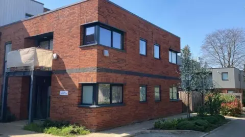 Brian Farmer/BBC Part of Greengates Court: A red-bricked building. Ten windows with grey frames are visible. A balcony over the entrance is covered with a tan-coloured material.