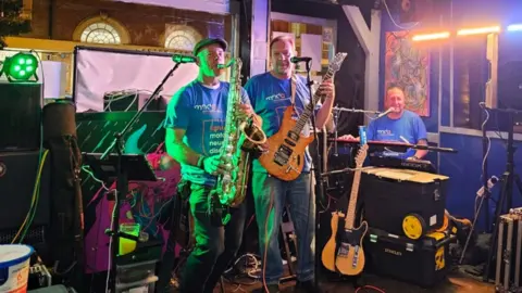 Christine Evans-Bradley Mark and Ian wearing blue 'fighting motor neurone disease' shirts whilst performing on stage. One is playing the saxophone and the other is playing a guitar.