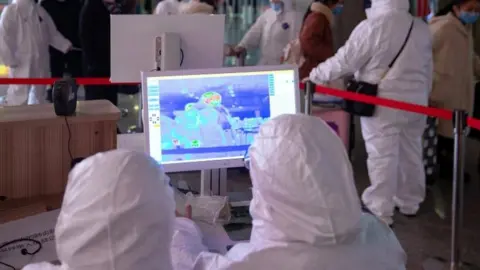 Reuters Workers in protective suits monitor a screen showing the thermal scan to check temperatures of passengers arriving at the Nanjing Railway Station, following the outbreak of a new coronavirus, during Chinese Lunar New Year holiday in Nanjing, China January 27, 2020.