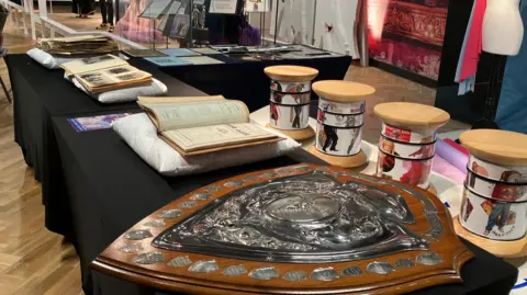 A series of items on a row of tables, which are covered with black tablecloths. They include a large wooden award shield with silver detailing, a book placed on a pillow, and a scrapbook with photographs. The tables are in a museum, with exhibits behind.