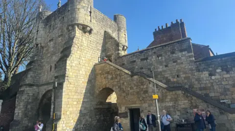 The Bootham Bar section of York's city walls, which includes a tower, two arches and a stone staircase. Tourists gather in front of it as a meeting point.