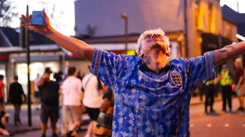 A man with dyed blonde hair and a blue-patterned England football shirt on holds his arms wide and throws his head back on a street in the evening sunlight as people walk past him in the background