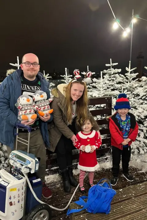 Family handout The Green family at a Christmas event in 2023. Evie is wearing a red Christmas outfit and is attached to a Berlin Heart machine. Her brother Theo is to her left, and their parents, David and Chloe, are next to them. Mr Green is holding two toy penguins. White Christmas trees are in the background of the photo.