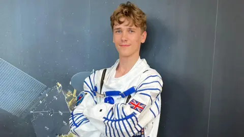 Natalia Maxim is wearing a space suit and posing behind a satellite print. 