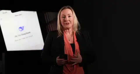 Zoe Kleinman holding a mobile phone, stood in front of a screen with the DeepSeek logo on (a purple whale)