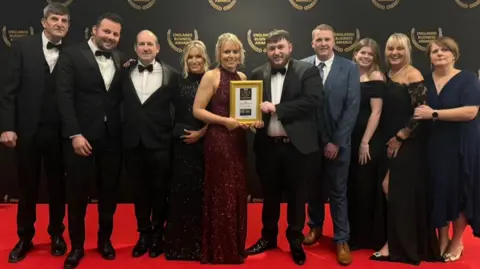 Steele's Family Bakery Ten people standing on a red carpet, posing for a photo. There are five men and five women. In the middle is Rachael who is blonde and wearing a red sparkly dress. She is holding a gold frame with an award inside it. Also holding the award is Joe, who is wearing a black and white suit and bow tie, and has brown hair and facial hair.
