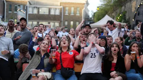 PA A group of England fans sitting in a fan zone. The are reacting to a moment in the game, with some people with their mouths open, others with hands on their heads, and others covering their mouths