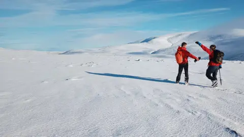 Benjamin Ross Two men wearing red jackets and black trousers, standing on a snowy mountaintop. The ground is completely white. A collection of hills in the background are also covered with snow. The sky above is light blue with streaks of white cloud.