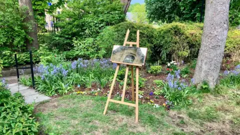 A wooden easel with a gold plaque propped up on it stands on a grassy section of a garden. Behind, a flowerbed with a variety of plants can be seen. To the left, a paved path and black handrail can be seen.