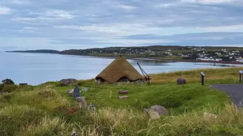 Gairloch Museum A visualisation of how the roundhouse would look at Gairloch Museum. The roundhouse has a conical, turf roof above stone walls. It is in an area of long grass and the sea is behind it.