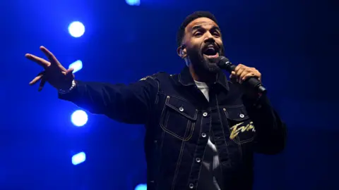 Getty Images Craig David is holding a microphone at a gig while singing. He's wearing a dark denim jacket, holding his hand out with bright stage lights behind him.