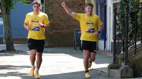 Helen Cowan The two brothers running along a street. They are wearing yellow t shirts and black shorts and orange running shoes. Euan is holding his right arm aloft in triumph.