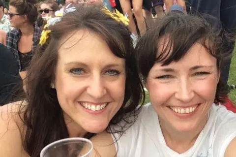Sharon Gorham Two women, Chelsea Booth and Sharon Gorham, are outdoors on a grassy area, at an event. They are smiling, one wearing a flower crown, the other wearing a white top. There are various people gathered in the background. 