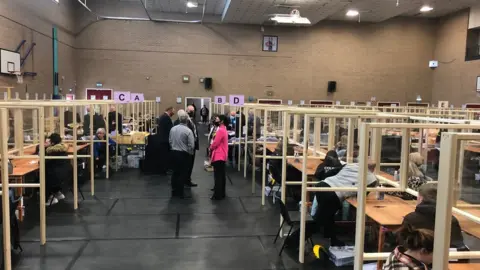 Candidates group in the centre of the room and counters sit at clear counting desks