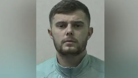 Northumbria Police Mugshot of Joyce-Scott. He has short dark hair and a beard and is wearing a light blue zip up jacket.