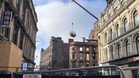 The facade of a Victorian building destroyed during a fire. The area is cordoned off with large barriers and a crane with a basket hovers above the ruins.